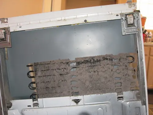 Close-up of dusty refrigerator condenser coils behind a kickplate.